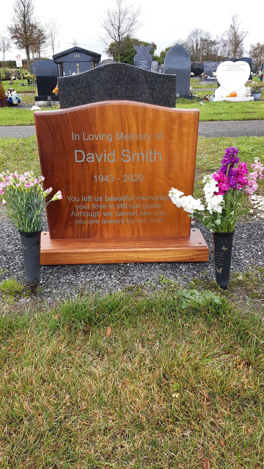 Iroko wooden headstone carved with a name and dates, standing in a cemetery